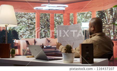 Entrepreneur analyzing architectural sketches and blueprints at his home, planning the layout and design of a new office to foster business development. Hunting for creative workspace. Camera B. 116437107