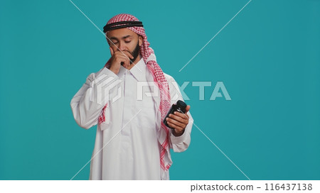 Muslim adult looks at jar of pills on camera, preparing to take painkillers or vitamins from bottle of medicine. Young person examining healthcare drugs in studio, medical treatment. 116437138