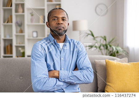 Confident man sitting on couch with arms crossed in modern living room. Background features bookshelf, plant, and clock adding to contemporary setting. Depicts confidence, determination relaxation. 116437274