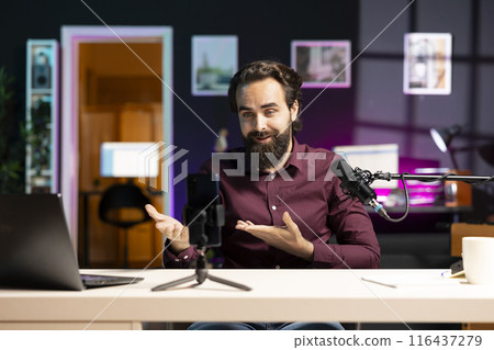 Smiling media star using smartphone on tripod to film vlog channel intro for online streaming platforms. Cheerful man greets viewers, presenting today video topic, recording with phone Smiling media star using smartphone on tripod to film vlog channel intro for online streaming platforms. Cheerful man greets viewers, presenting today video topic, recording with phone 116437279