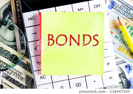 Business concept. BONDS word on a yellow sticker on a calculator against the background of American dollars Business concept. BONDS word on a yellow sticker on a calculator against the background of American dollars 116447503