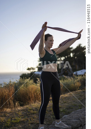 A woman is exercising outdoors, holding a purple band 116448515