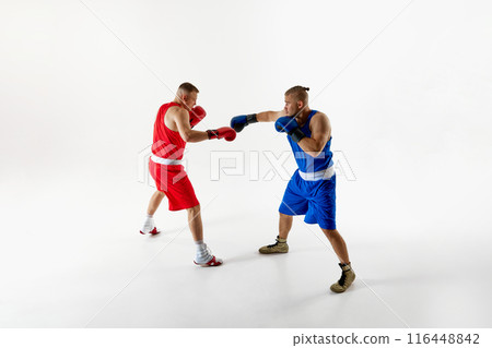 Two determined boxers in blue and red attire in action refining their techniques during intense training session against white studio background. Two determined boxers in blue and red attire in action refining their techniques during intense training session against white studio background. 116448842