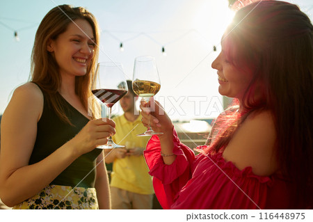 Photo of lively toast among friends, two beautiful women. Their faces lit up with smiles and drinks sparkling in sun. 116448975