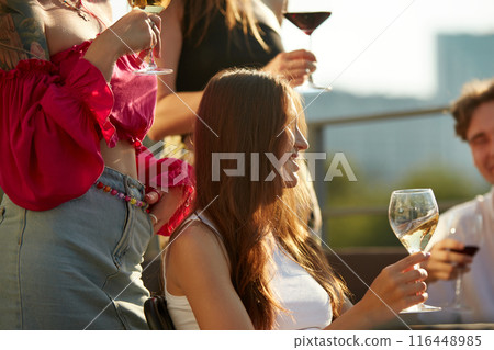 Friends, young men and women, toast to good times, their glasses meeting in center as they bask in sunlight. 116448985