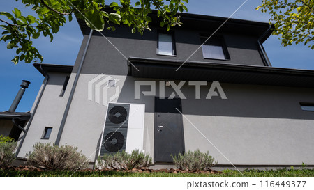 Positioned by the wall of a modern cottage, the outdoor unit of a heat pump is visible. Positioned by the wall of a modern cottage, the outdoor unit of a heat pump is visible. 116449377