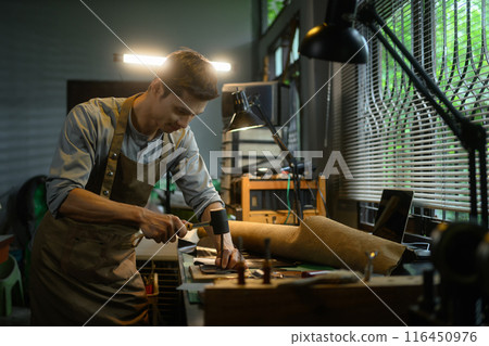 Handsome male tailor working with leather using hammer in his workshop. Small business and handmade concept Handsome male tailor working with leather using hammer in his workshop. Small business and handmade concept 116450976
