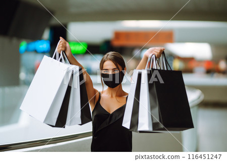 Young woman in protective black medical mask on her face with shopping bags in the mall.Covid-2019. Young woman in protective black medical mask on her face with shopping bags in the mall.Covid-2019. 116451247