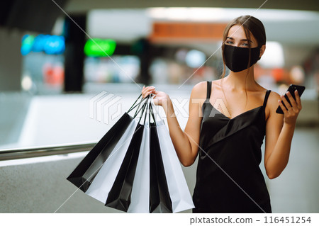 Young woman in protective black medical mask with shopping bags talking on a mobile phone in the mall. 116451254