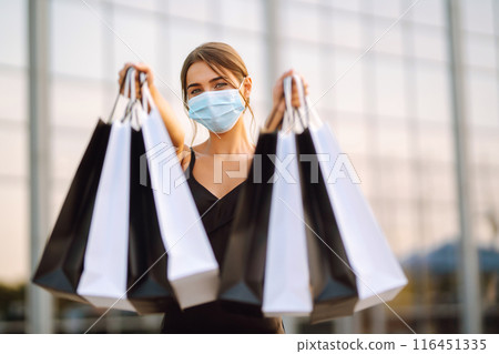 Beautiful woman in protective sterile medical mask with shopping bags near shopping center.Covid- 2019. Beautiful woman in protective sterile medical mask with shopping bags near shopping center.Covid- 2019. 116451335