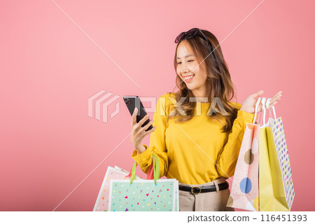 Portrait Asian happy beautiful young woman teen shopper smiling standing excited holding online shopping bags online colorful on mobile phone or smartphone studio shot isolated on pink background 116451393