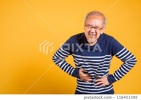Portrait Asian old man with glasses holds hands on abdomen he stomach ache studio shot isolated yellow background, senior man pensioner feeling pain abdominal pain, hepatitis painful 116451395