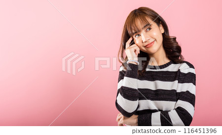 Portrait of a contemplative Asian woman, standing with her chin in her hand, thinking about a question. Studio shot on pink with ample copy space, showing her intelligent look. 116451396