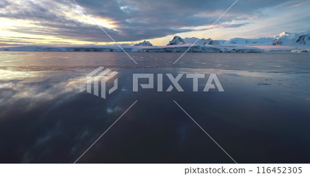 Low angle flight over polar icy ocean in Antarctica. Snow-covered mountains range under colorful sunset clouds sky. Tranquil scene icy landscape, wild nature background. South Pole travel, exploration Low angle flight over polar icy ocean in Antarctica. Snow-covered mountains range under colorful sunset clouds sky. Tranquil scene icy landscape, wild nature background. South Pole travel, exploration 116452305