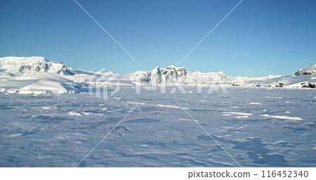 Aerial fly over winter mountain landscape in Antarctica. Polar frozen ocean landscape covered by snow under blue sunny sky. Discover the beauty of South Pole. Antarctica travel exploration background 116452340