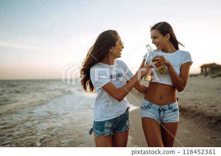 Beautiful two girls with glasses is holding a bottle with beer in her hands at the beach.. Summer lifestyle. Beautiful two girls with glasses is holding a bottle with beer in her hands at the beach.. Summer lifestyle. 116452381