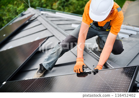 Worker building photovoltaic solar panel system on rooftop of house. Man engineer in helmets and gloves installing solar module with help of hex key outdoors. Alternative energy generation concept. Worker building photovoltaic solar panel system on rooftop of house. Man engineer in helmets and gloves installing solar module with help of hex key outdoors. Alternative energy generation concept. 116452402