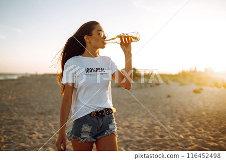 Pretty girl drinking beer at the beach at sunset. Young woman enjoying on beach holiday. Summertime 116452498