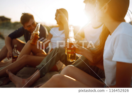 Young people sitting together at beach, drinking beer. Group of friends cheers with beers at the beach. 116452926