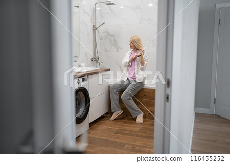 Confident blond woman combs her long hair in modern bathroom, exuding beauty and selfassurance 116455252