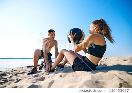 Man helping woman with hard press exercises with medicine ball on sandy beach under clear blue sky. Intensive workout 116455957