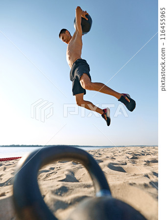 Shirtless man in black shorts jumping with medicine ball on sandy beach, with clear blue sky and distant water in the background. Shirtless man in black shorts jumping with medicine ball on sandy beach, with clear blue sky and distant water in the background. 116455965