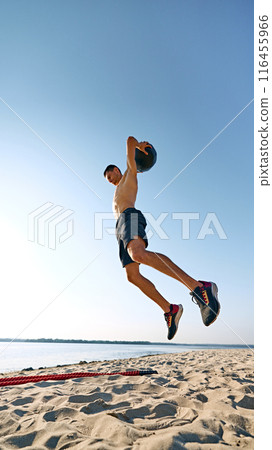 Shirtless man in black shorts jumping with medicine ball on sandy beach, with clear blue sky and distant water in the background. 116455966