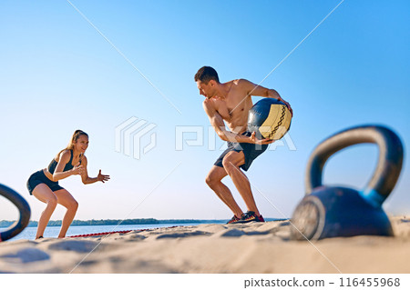 Man and woman exercising on sandy beach with medicine ball, with under a clear blue sky. Intensity functional training 116455968