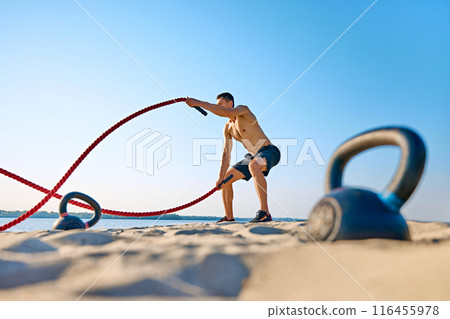 Muscular shirtless man in black shorts training with battle ropes on sandy beach under blue clear sky, with kettle bell lying in sand. 116455978