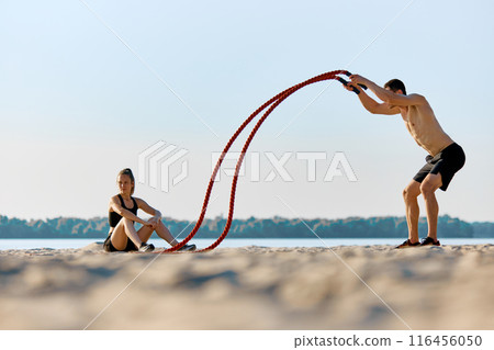 Shirtless man in black shorts, with muscular relief body training with battle ropes on sandy beach by the river, while woman sitting on sand and watching exercises Shirtless man in black shorts, with muscular relief body training with battle ropes on sandy beach by the river, while woman sitting on sand and watching exercises 116456050