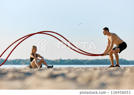 Shirtless man in black shorts, with muscular relief body training with battle ropes on sandy beach by the river, while woman sitting on sand and watching exercises 116456051