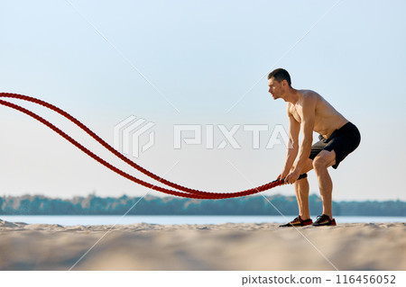 Shirtless man with muscular fit body training on sunny beach with battle ropes, hard functional open air training session. 116456052