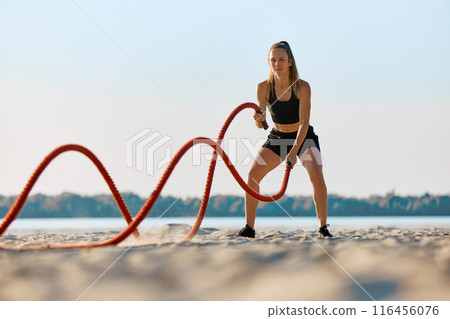 Athletic woman in black sportswear exercising with red battle ropes on a sandy beach, with water and trees in background under clear sky. 116456076