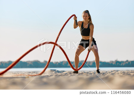 Woman in black sportswear exercising with red battle ropes on a sandy beach, with water and trees in background under clear sky. Woman in black sportswear exercising with red battle ropes on a sandy beach, with water and trees in background under clear sky. 116456080