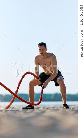 Athlete performing battle rope exercises on sandy beach, shirtless muscular man in n black shorts doing intensity functional training outdoors 116456084