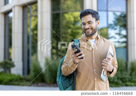 University student standing outside modern building smiling while looking at phone. Student carrying backpack and folder, representing education, technology, and youth lifestyle on campus. University student standing outside modern building smiling while looking at phone. Student carrying backpack and folder, representing education, technology, and youth lifestyle on campus. 116456668