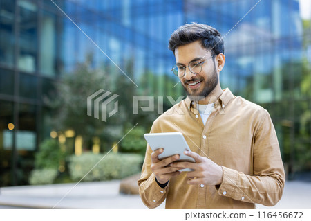 Young man smiling while using tablet outside modern office building. Casual attire, urban setting, and contemporary design. Represents technology, business, and communication. Happy expression 116456672