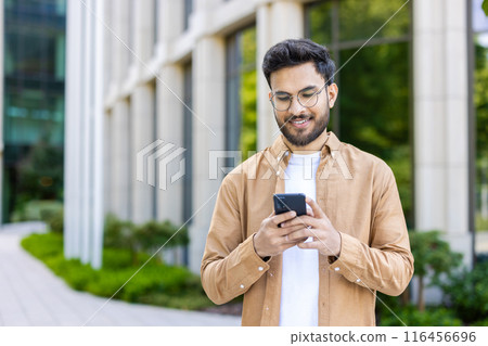 Smiling young man wearing glasses using smartphone outside modern office building. Casually dressed man engaging with social media or business apps phone, bright environment representing technology 116456696