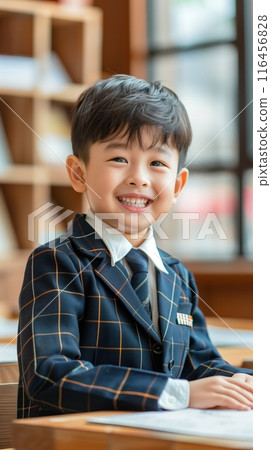 Seven year old Asian boy wears blue orange plaid suit with white collar and black tie. Smiles at camera in classroom with bookshelf full of books suggesting knowledge learning. Back to school theme. 116456828