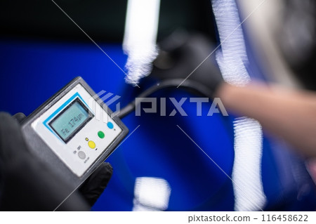 Image shows a person measuring the paint thickness on a blue car  116458622
