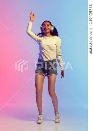 Full-length image of young girl in shorts and t-shirt listening to music in headphones and dancing with joy against gradient pink blue background in neon light Full-length image of young girl in shorts and t-shirt listening to music in headphones and dancing with joy against gradient pink blue background in neon light 116459758