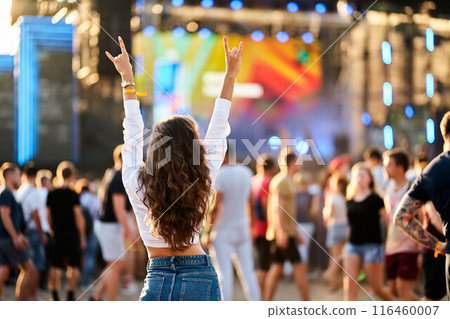Back view of young woman with raised hands enjoys live music at sunny beach festival. Happy crowd around stage, summer event vibe. Fan cheers band, fun outdoor concert. Casual denim, festival fashion. 116460007