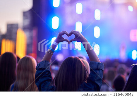 Back view of person forming heart shape with hands at music fest, surrounded by cheering crowd, vibrant stage lights blur in background, festive event atmosphere captures moment of joy at live concert Back view of person forming heart shape with hands at music fest, surrounded by cheering crowd, vibrant stage lights blur in background, festive event atmosphere captures moment of joy at live concert 116460042