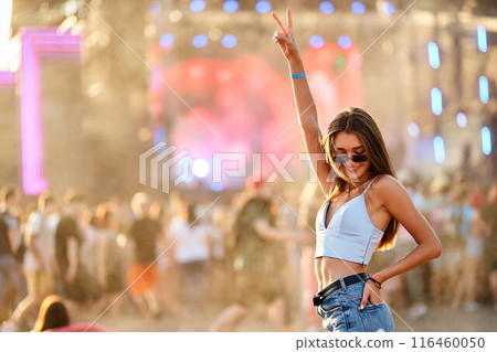 Smiling young woman enjoys summer music festival on beach, dancing among crowd. Hand up with peace sign, trendy sunglasses and casual festival wear. Sunset lights add warm, vibrant feel to scene. 116460050