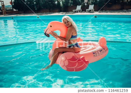A woman is sitting on a pink inflatable flamingo in a pool. The flamingo is pink and has a blue stripe. The woman is smiling and she is enjoying herself. Concept of fun and relaxation. A woman is sitting on a pink inflatable flamingo in a pool. The flamingo is pink and has a blue stripe. The woman is smiling and she is enjoying herself. Concept of fun and relaxation. 116461259