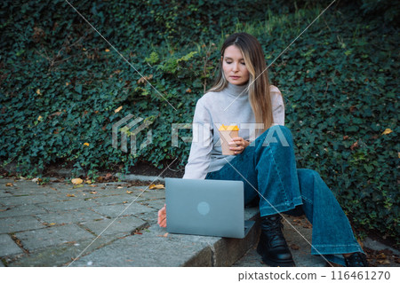 A woman is sitting on a stone ledge with a laptop and a cup of coffee. She is working or studying, as she is focused on her laptop. Concept of productivity and concentration. A woman is sitting on a stone ledge with a laptop and a cup of coffee. She is working or studying, as she is focused on her laptop. Concept of productivity and concentration. 116461270