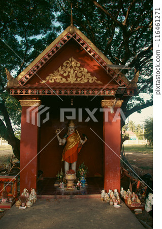 A spirit house or tope taveda with a deity statue, adorned with red and gold decorations, surrounded by offerings for the neak ta or guardian spirit set amidst trees in Kampot Cambodia A spirit house or tope taveda with a deity statue, adorned with red and gold decorations, surrounded by offerings for the neak ta or guardian spirit set amidst trees in Kampot Cambodia 116461271