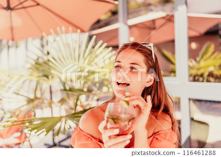 A woman in an orange dress is sitting in a chair with a drink in front of her. She is looking at the camera and she is enjoying her drink. The scene is set in a patio or outdoor area. 116461288