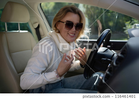 A blonde woman in a white sweater and jeans is driving. Happy woman sitting in a car with a white interior. 116461401