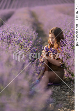 Girl is sitting in a field of purple flowers. She is holding a basket of flowers and smiling. Scene is peaceful and serene, as the girl is surrounded by the beauty of nature. 116461428
